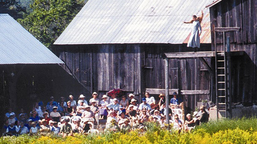 A seated audience looks up as an actor performs on a raised stage next to a barn.