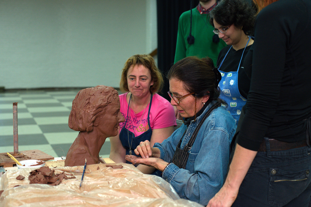 A woman sits working on a sculpture of a bust while a group of people watch.