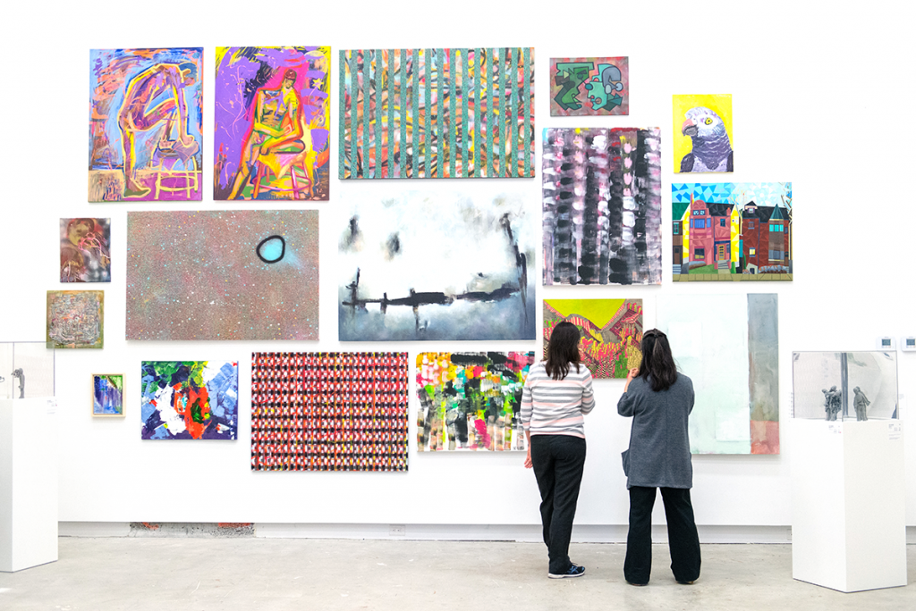 Two people stand facing a wall of full of hanging visual artworks in a gallery.