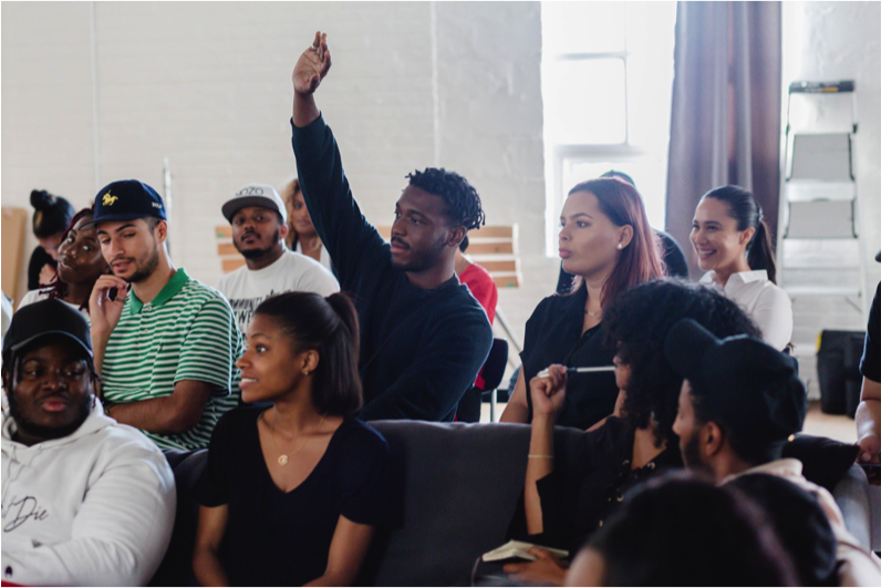 The Remix Project participants sit in a class while one of the students raises his hand.