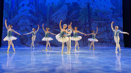 Ballet dancers perform on a stage with a blue backdrop displaying trees covered in snow.