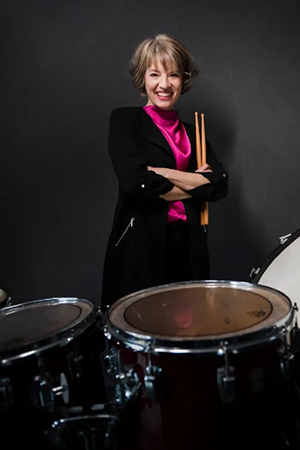 Person standing confidently behind a drum set, holding drumsticks with arms crossed against a dark background.