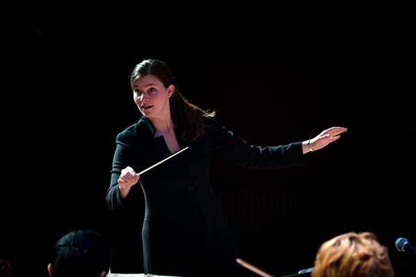Person in a dark outfit conducting an ensemble on stage, holding a baton with one arm extended