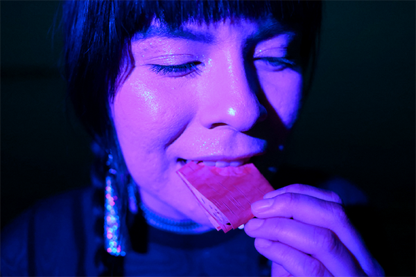 Close-up of a person holding a piece of birch bark near the mouth, appearing to bite it. The scene is illuminated, creating a glowing effect.