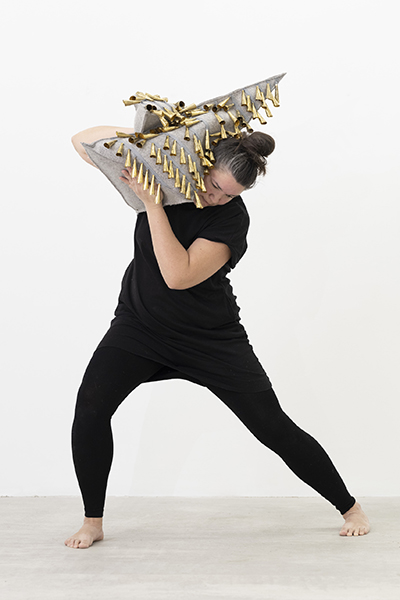 A person stands in a wide stance, holding a large triangular sculptural piece made of gray material adorned with rows of golden cone-shaped jingles. The object is positioned diagonally across the upper body, creating a dynamic sense of movement.