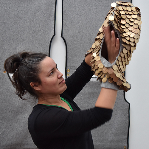 Une personne tient une pièce sculpturale composée de pièces dorées superposées, formant une surface texturée semblable à des écailles. La pièce se courbe vers le haut et l’extérieur à partir des mains, créant une forme allongée et creuse.