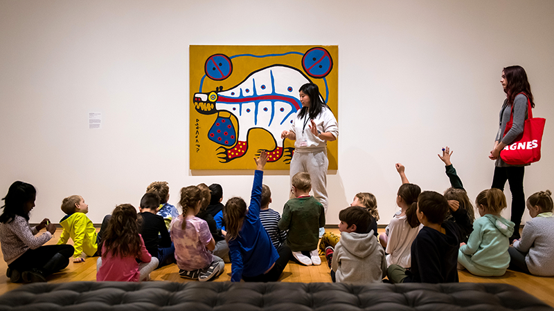 Une éducatrice est debout devant un groupe d’enfants assis par terre qui regardent une œuvre d’art visuel représentant un ours. Certains enfants lèvent la main.