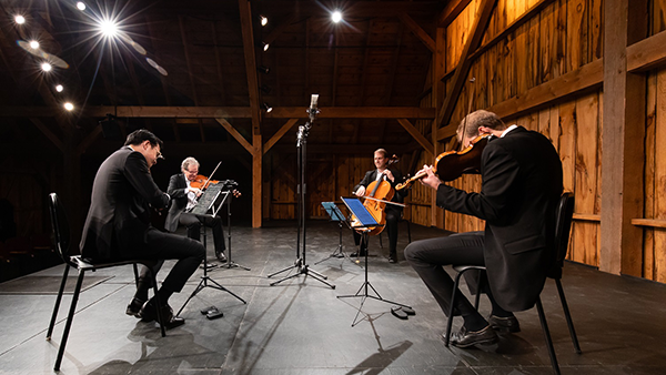 New Orford String Quartet at Westben Two seated men sit facing each other on a stage playing string instruments.