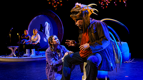 A girl crouches on a stage looking up at a seated man dressed as a porcupine. Two women sit in the background looking towards the two characters in the foreground. A girl crouches on a stage looking up at a seated man dressed as a porcupine. Two women sit in the background looking towards the two characters in the foreground.