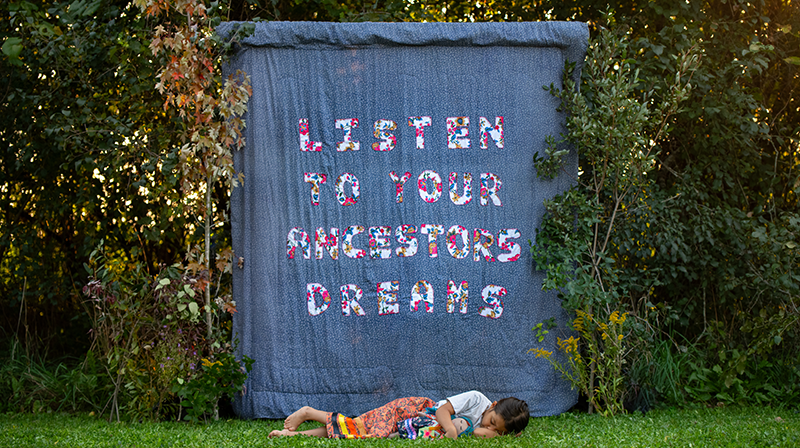 In this horizontal photograph, a child sleeps on the grass cradling a doll. Both she and the doll wear ribbon skirts. Hanging behind them, framed by trees, is a blue blanket with letters made of floral fabric. They read: "Listen to your ancestors’ dreams".