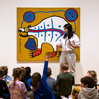 An educator stands in front of a crowd of kids sitting on the ground as they look up at an Indigenous visual artwork of a bear. Some of the children are raising their hands.