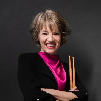 Person standing confidently behind a drum set, holding drumsticks with arms crossed against a dark background.
