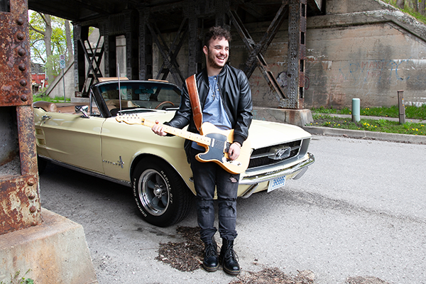 Evan Rotella tient une guitare devant une Ford Mustang décapotable jaune des années 1960 sous un pont.