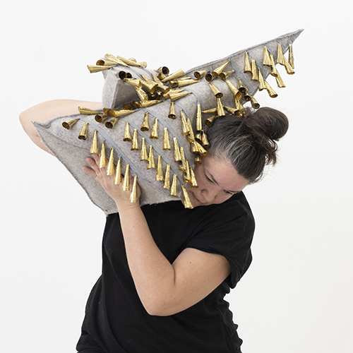 A person stands in a wide stance, holding a large triangular sculptural piece made of gray material adorned with rows of golden cone-shaped jingles. The object is positioned diagonally across the upper body, creating a dynamic sense of movement.