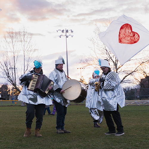 Performers dressed in mostly white with black bottoms march around on grass in a park. Three people play instruments – the accordion, a bass drum and a saxophone. Another walks behind waving a large white flag with a red heart. 
