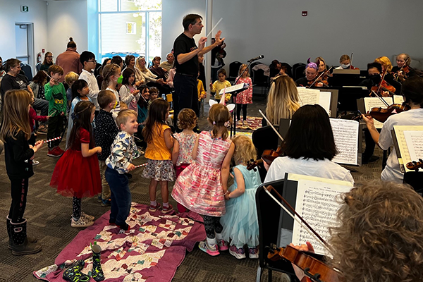 A standing man conducts seated musicians. He is surrounded by attentive small children who hold smaller striped batons.