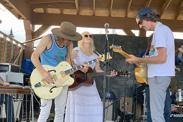 Three people standing on an outdoor stage, behind a microphone, all playing guitars.
